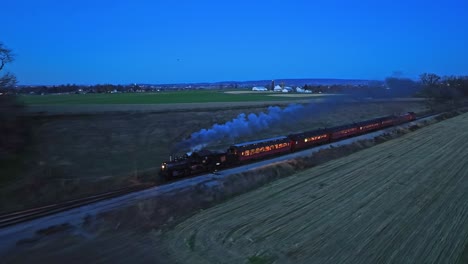 Una-Vista-Nocturna-De-Drones-De-Un-Tren-De-Pasajeros-De-Vapor-Que-Se-Acerca-En-Una-Sola-Vía-Que-Viaja-A-Través-De-Tierras-De-Cultivo-Y-Ve-Las-Luces-En-Los-Vagones