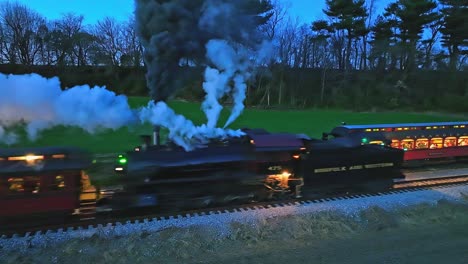 Un-Dron-Ligeramente-Por-Delante-Y-Una-Vista-Nocturna-Paralela-De-Un-Tren-De-Pasajeros-De-Vapor-Se-Detuvo-Cuando-Otro-Tren-De-Vapor-Pasa-Soplando-Mucho-Humo-Viendo-Las-Luces-En-Los-Vagones