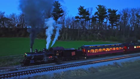 Un-Dron-Ligeramente-Por-Delante-Y-Una-Vista-Nocturna-Paralela-De-Un-Tren-De-Pasajeros-De-Vapor-Detenido-En-Tierras-De-Cultivo-Que-Sopla-Mucho-Humo-Al-Ver-Las-Luces-En-Los-Vagones