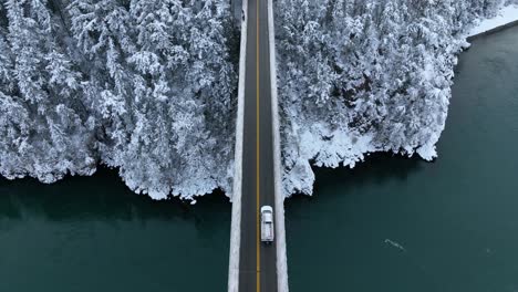 Aerial-view-of-a-truck-driving-across-a-bridge-and-entering-a-snow-covered-forest