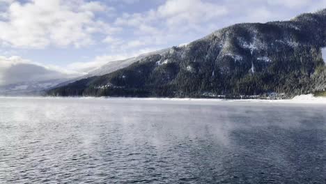 Steam-rising-off-the-top-of-Upper-Arrow-Lake-with-a-landscape-view-of-the-mountains-with-alpine-trees-covered-in-snow
