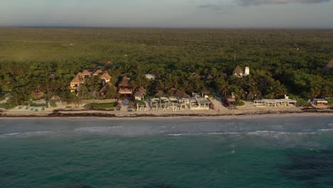 Aerial-backwards-shot-of-expensive-five-star-villas-with-private-sandy-beach-and-palm-trees-in-front-of-Caribbean-Sea-during-sunset-in-Tulum,Mexico---Beautiful-green-growing-landscape-in-background