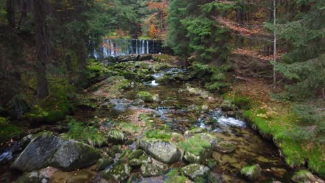 Fliegen-über-Einem-Flussbett-Mit-Felsbrocken-Und-Einem-Künstlichen-Wasserfall-In-Einem-Herbstlichen-Mischwald