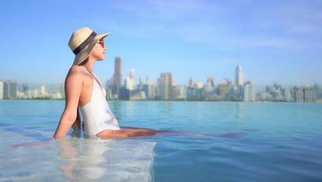 Sexy-Asian-Woman-in-Swimsuit-Sitting-on-Rooftop-Infinity-Pool-Edge-With-Stunning-View-of-Modern-Metropolis-in-Background