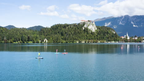 AERIAL---People-on-paddleboards-in-Lake-Bled,-Julian-Alps,-Slovenia,-lowering-shot