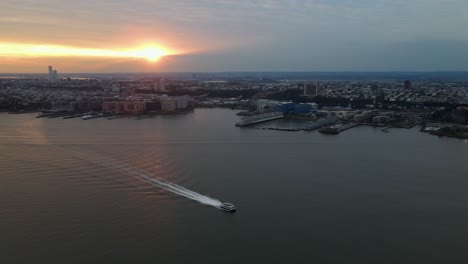 Aerial-view-of-a-ferry-on-Hudson-river,-sunset-Jersey-city-background---pan,-drone-shot
