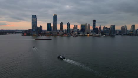 Aerial-view-of-ferries-in-front-of-the-Jersey-city-skyline,-in-cloudy-sunset-in-NY,-USA---rising,-drone-shot