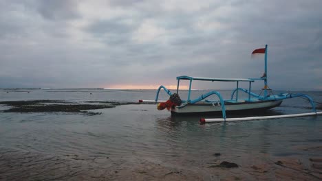 Seitlicher-Drohnenschuss-Eines-Indonesischen-Fischerbootes-Auf-Dem-Meer-Mit-Dem-Sonnenuntergang-Im-Hintergrund-Am-Strand-Von-Sanur,-Bali-Indonesien-In-4k