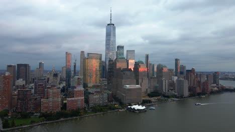 Aerial-drone-view-towards-sunlit-skyscrapers-in-lower-Manhattan,-dark,-cloudy-sunset-in-New-York,-USA
