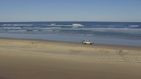 Beautiful-tracking-shot-of-a-white-four-wheel-drive-truck-driving-along-a-deserted-Queensland-beach-under-a-stunningly-clear-sky-and-gentle-rolling-surf