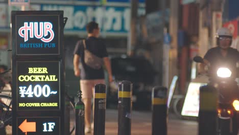 Lighted-Sign-Of-A-British-Pub-With-People-Walking-And-Driving-A-Motorcycle-In-The-Street-At-Night-In-Kamata,-Tokyo,-Japan