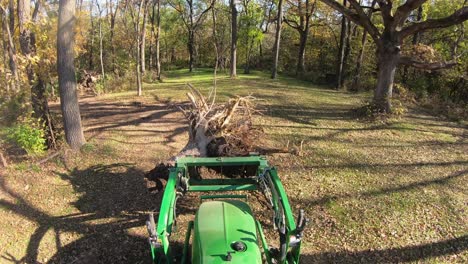 High-angle-point-of-view-on-small-green-tractor-using-lift-forks-to-move-a-tree-stump-thru-a-clearing-in-the-woods-in-autumn