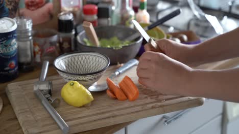 Hands-of-woman-cutting-carrot-on-cutting-board-next-to-lemon,-avocado-halves-and-garlic-press-in-a-messy-kitchen