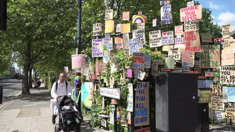 Un-Hombre-Con-Un-Niño-En-Un-Cochecito-Pasa-Por-Un-Enorme-Muro-Conmemorativo-De-Carteles-De-Cartón-Que-Muestran-El-Apoyo-Del-Personal-De-Atención-Médica-Durante-El-Brote-De-Coronavirus-En-El-Borde-De-Un-Parque-En-El-Extremo-Este-De-Londres