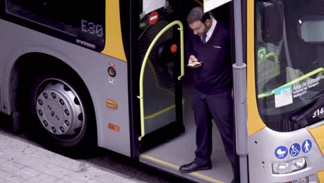 Covid-19:-Cierre-Manual-De-Un-Conductor-conductor-Revisando-Su-Móvil-Parado-En-La-Puerta-Vacía-Del-Autobús