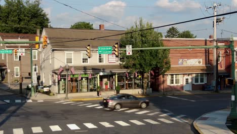 American-flags-at-business-store,-storefront-at-intersection-of-small-town-in-United-States,-car-traffic-passes-by-during-magic-hour,-aerial-push-in-establishing-shot