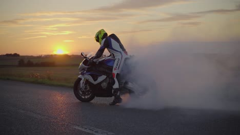 Tiro-De-Cámara-Lenta-De-Ancho-Medio-De-Un-Motociclista-Con-Traje-De-Cuerpo-Completo-Y-Casco-Realizando-Un-Agotamiento-Con-Una-Superbike-Blanca-Durante-La-Puesta-De-Sol