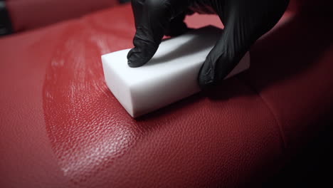 Tracking-close-up-4k-shot-of-a-male-hand-in-a-black-rubber-glove-cleaning-a-red-leather-car-seat-using-a-white-sponge-and-a-car-polish-spray