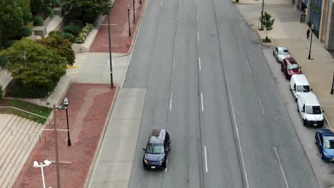 Aerial-tilt-down-tracks-a-blue-SUV-on-urban-city-street-in-United-States,-comes-to-stop-at-intersection,-skyscrapers-in-downtown