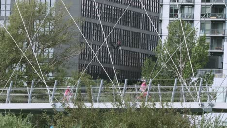 Young-girls-bicycle-on-Calgary-suspension-bridge,-window-washer-beyond