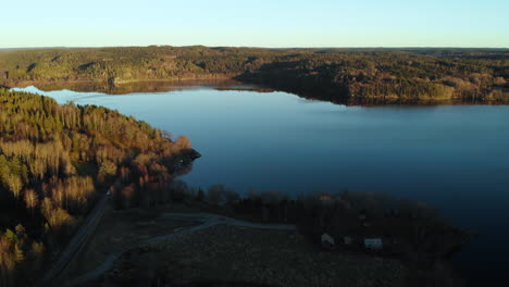 Aerial-wide-shot-view-over-a-blue-lake-surrounded-by-a-green-forest-on-a-sunny-day,-drone-flying-backward