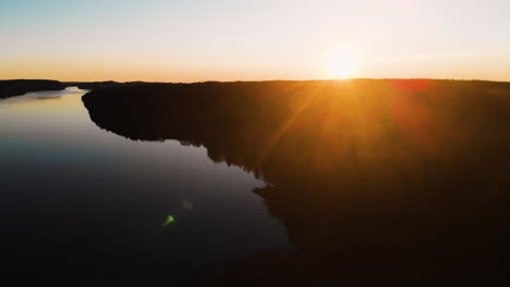Sunset-over-Black-Silhouetted-Forest-Next-Calm-Reflecting-Lake-AERIAL