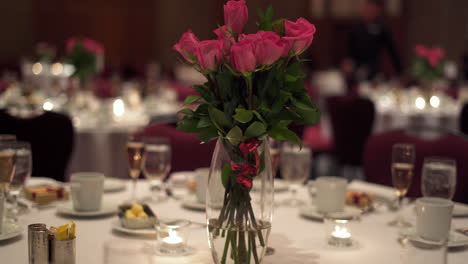 Long-stemmed-pink-roses-in-glass-vase-on-set-table-with-white-tablecloth-in-banquet-hall-for-special-occasion-wedding