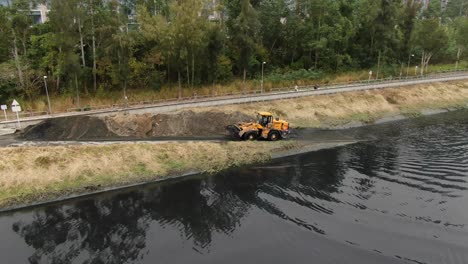 Gelber-Traktor,-Der-Schlamm-Aus-Einem-Kanal-Unter-Einer-Brücke-Schöpft-Und-Reinigt,-Luftbild