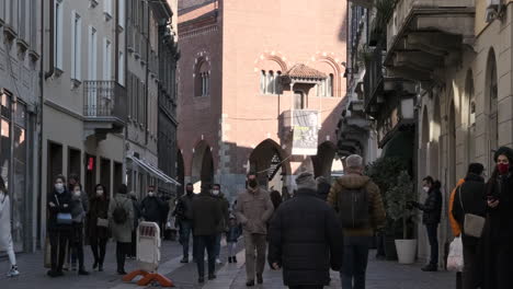 Menschen,-Die-Medizinische-Masken-Für-Die-Covid-19-pandemie-Tragen-Und-An-Einem-Sonnigen-Januartag-In-Monza,-Norditalien,-Auf-Der-Stadtstraße-Spazieren-Gehen---Mittlere-Aufnahme