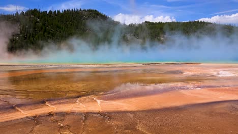 Static-view-of-geothermal-geyser-and-hot-spring-with-steam-cloud-rising-and-mountains-in-the-background-at-Grand-Prismatic-Spring,-Yellowstone-National-Park,-Wyoming,-USA