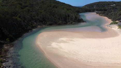 Revealing-drone-shot-of-the-Korogoro-Creek-and-tropical-mountains-with-wind-blowing-sand-across-a-sand-bar-at-Hat-Head-New-South-Wales,-Australia