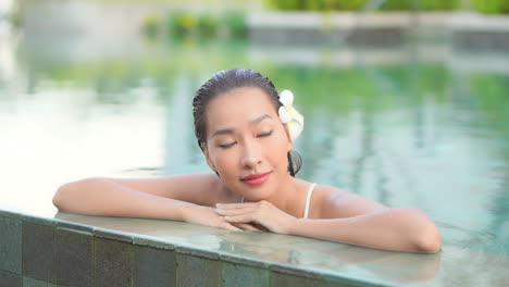 Slow-motion-view-of-attractive-Asian-woman-with-flower-behind-ear-standing-in-swimming-pool-resting-head-on-hands-and-looking-around