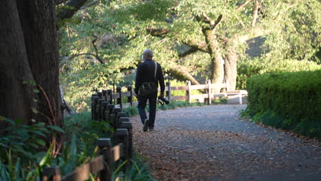 Back-View-Of-An-Old-Man-Photographer-Carrying-His-DSLR-Camera-In-A-Nature-Park-In-Tokyo,-Japan---Medium-Shot,-Slow-Motion