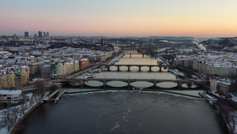 Prague-Czech-Republic-at-cold-winter-evening,-aerial-view,-bridges-above-Vltava-river,-snow-capped-buildings-and-clear-sky-after-sunset,-drone-shot