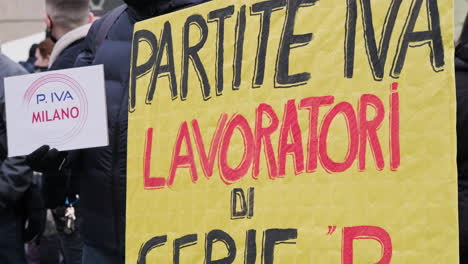 Un-Hombre-Sostiene-Un-Cartel-Amarillo-De-Protesta-Contra-El-Gobierno-Italiano-Durante-Una-Manifestación-En-La-Plaza-De-Milán,-Italia