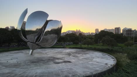 Dolly-in-flying-over-Floralis-Generica-steel-sculpture-leading-to-Recoleta-buildings-at-golden-hour,-Buenos-Aires