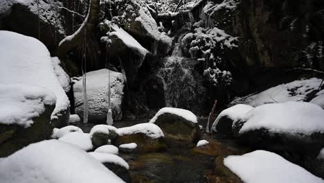 Clear-slow-streaming-creek-between-brown-rocks-and-stones-covered-with-white-snow-and-ice-with-a-waterfall-on-the-background