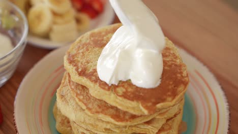 Stack-of-Crepes-on-Plate,-Apllying-Sweet-Greek-Yoghurt-on-Top,-Fruits-in-Background