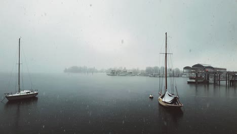 Snow-falling-over-Maggiore-lake-and-anchored-sailing-boat-and-ferry,-Italy