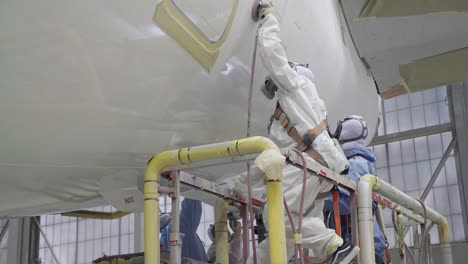 A-slow-motion-shot-of-two-blue-collar-hispanic-men-in-the-aviation-industry-use-a-scissor-lift-to-spray-a-top-coat-of-gloss-paint-on-the-side-of-a-commercial-airplane-in-a-busy-industrial-hangar