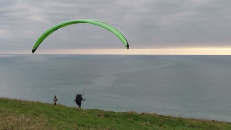 Ein-Mann,-Der-Versucht,-Mit-Seinem-Gleitschirm-Einen-Wind-Zu-Fangen,-Während-Eine-Frau-An-Einem-Bewölkten-Tag-Mit-Sonnenuntergangssonne-über-Dem-Horizont-Am-Rand-Der-Klippe-Mit-Blick-Auf-Den-Atlantik-Zuschaut