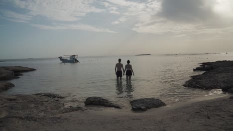 Couple-walking-out-into-the-sea-at-calm,-beautiful-sunset