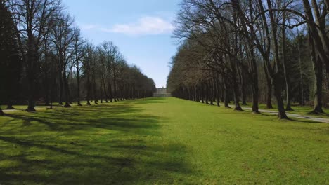 Beautiful-low-flight-aerial-along-a-tree-alley-leading-up-to-Bavaria's-famous-castle-island-Herrenchiemsee-and-its-royal-park-at-lake-Chiemsee-in-the-rural-countryside-with-a-beautiful-blue-sky