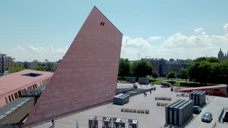 Vuelo-Aéreo-Sobre-El-Museo-De-Gdansk-De-La-Segunda-Guerra-Mundial-Cuando-Los-Turistas-Caminan-Alrededor-Del-Edificio,-Banderas-De-Polonia-Y-Ciudad-Ondeando-En-Astas-De-Bandera