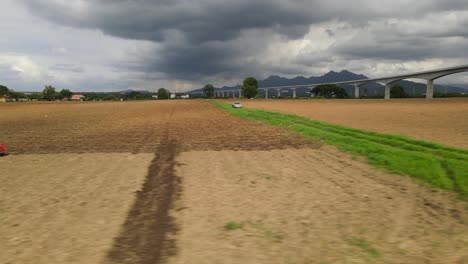 Imágenes-Aéreas-Deslizándose-Hacia-La-Derecha-Que-Revelan-Un-Tractor-Labrando-Una-Tierra-De-Cultivo-Que-También-Muestra-Un-Tren-Elevado-De-Alta-Velocidad-Y-Un-Hermoso-Paisaje-En-El-Horizonte