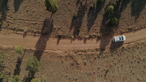 Top-down-rising-aerial-as-white-car-passes-under-drone-on-dusty-dirt-road,-4K