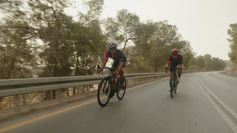 two-road-cycelist-professionals-riding-in-the-forest's-empty-road-and-the-sun-flares-to-the-camera