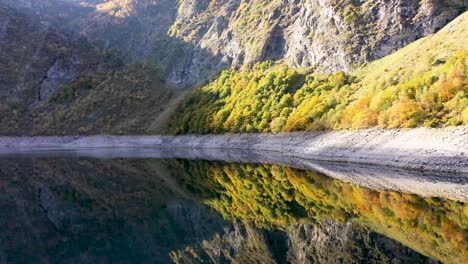 Lac-d'Oô-reflecting-artificial-lake-in-the-French-Pyrenees-with-view-of-the-Oo-waterfall,-Aerial-pan-left-reveal-shot