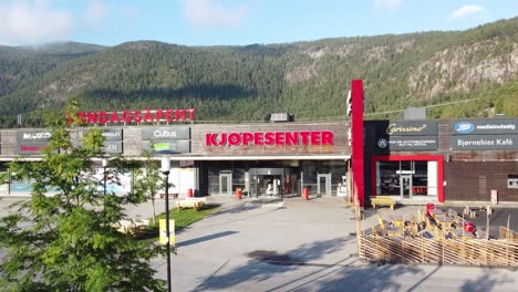 Bjorneparken-shopping-mall-in-Flaa-Norway---Sunny-day-ascending-aerial-behind-tree---Revealing-full-front-exterior-of-mall-with-lush-mountain-background