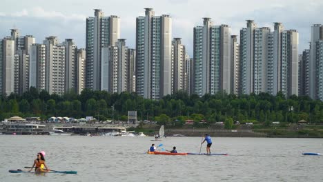 Active-Tourists-On-Paddle-Boarding-Sports-At-Han-River-With-Jamsil-District-Cityscape-In-Seoul,-South-Korea
